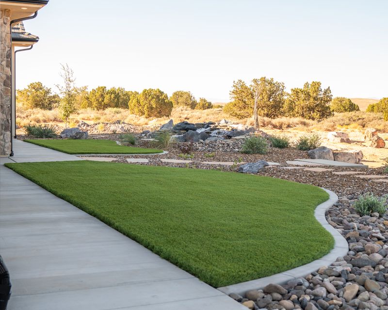 Curved turf lawn with concrete curbing along a walkway, with river rock, desert plantings, and a waterfall in the background