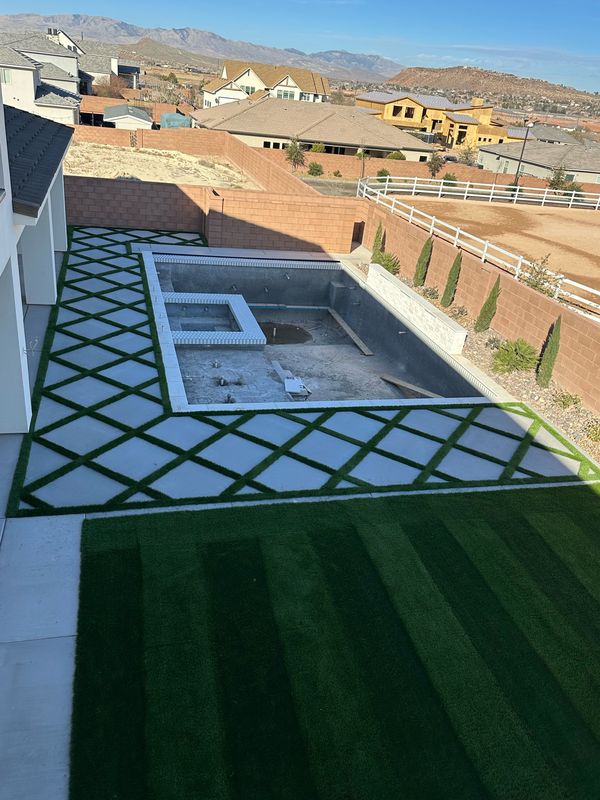 Large diamond-pattern concrete pavers with turf strips surrounding a pool under construction, with a striped turf lawn in front