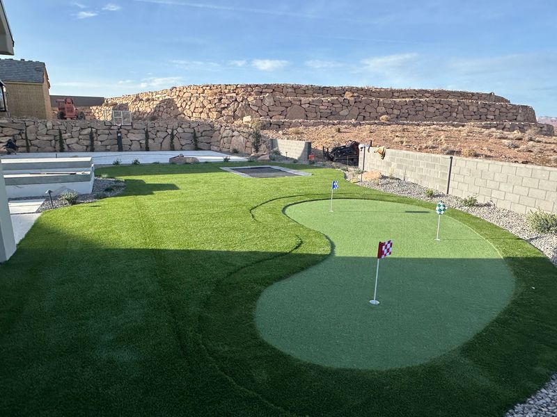 Putting green with three flags in a turf yard with boulder retaining walls, an in-ground trampoline, and a basketball hoop behind