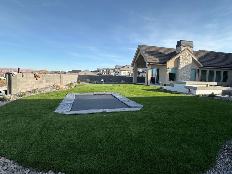 Large turf backyard with an in-ground trampoline and a putting green in the background, next to a home with a stone chimney