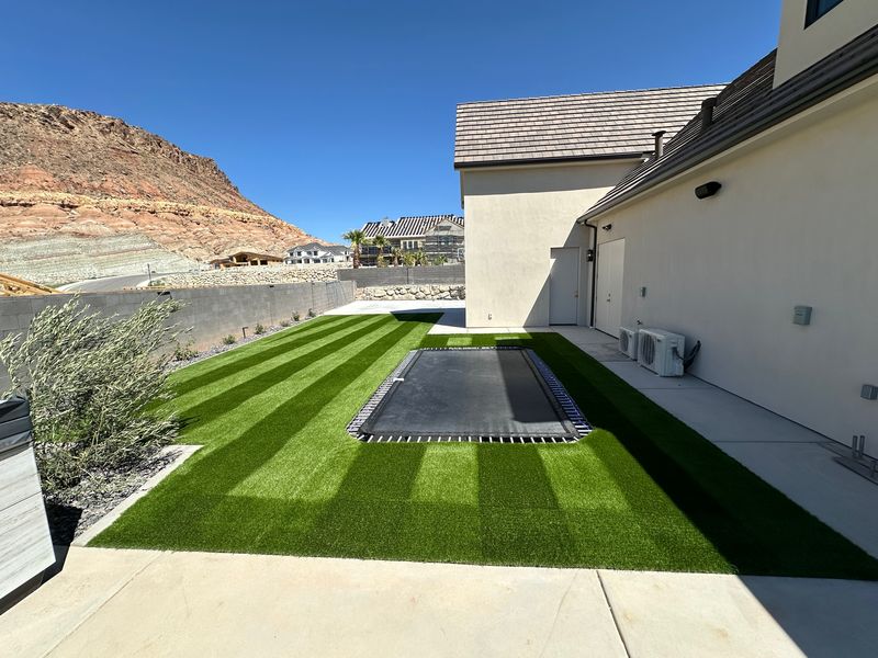 Striped artificial turf side yard with an in-ground trampoline and a red rock cliff in the background